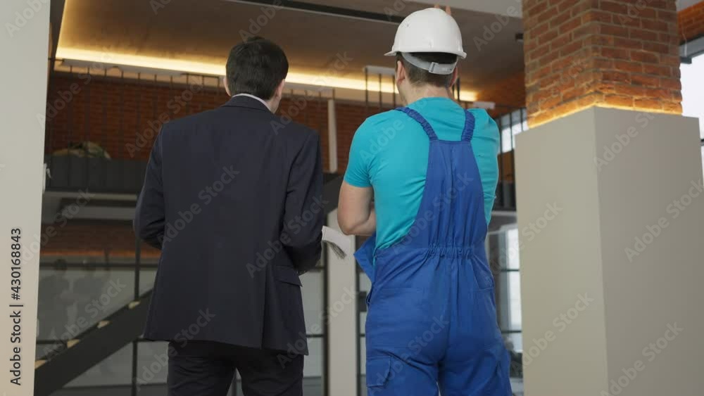 Back view of professional service man and premises owner talking discussing renovation indoors. Confident Caucasian man in hard hat and uniform showing repair to client in suit. Industrial concept Back view of professional service man and premises owner talking discussing renovation indoors. Confident Caucasian man in hard hat and uniform showing repair to client in suit. Industrial concept