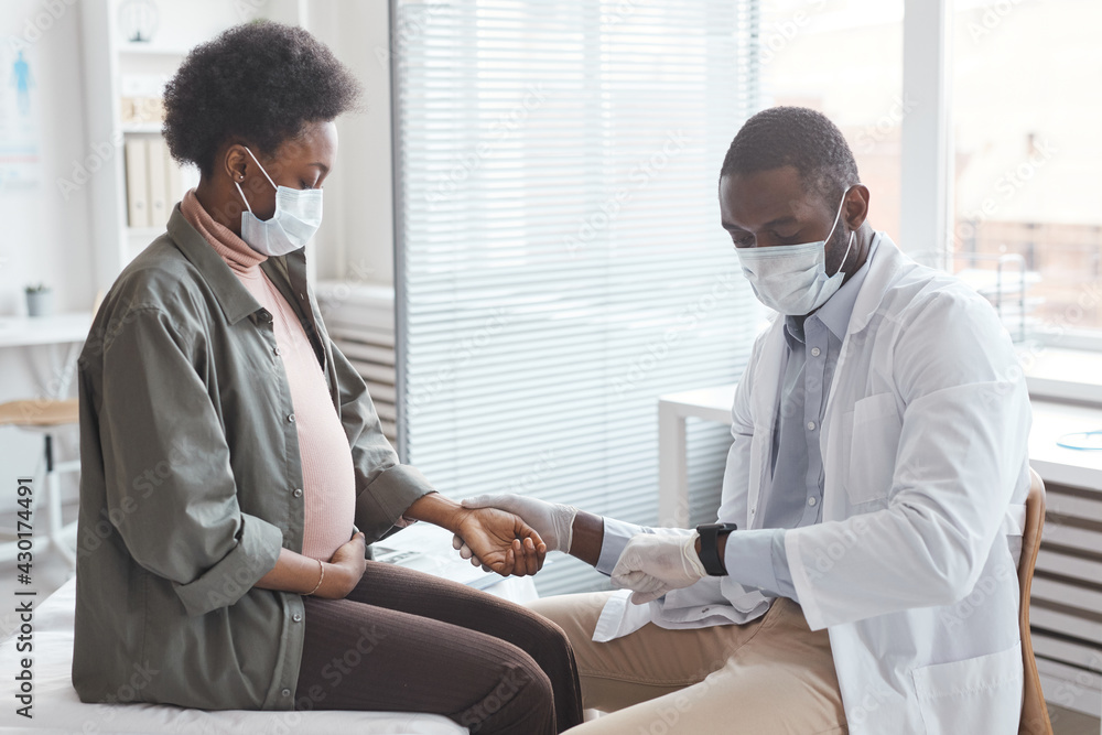 Obraz premium African young man in mask measuring the pulse of pregnant woman and caring about her health at hospital
