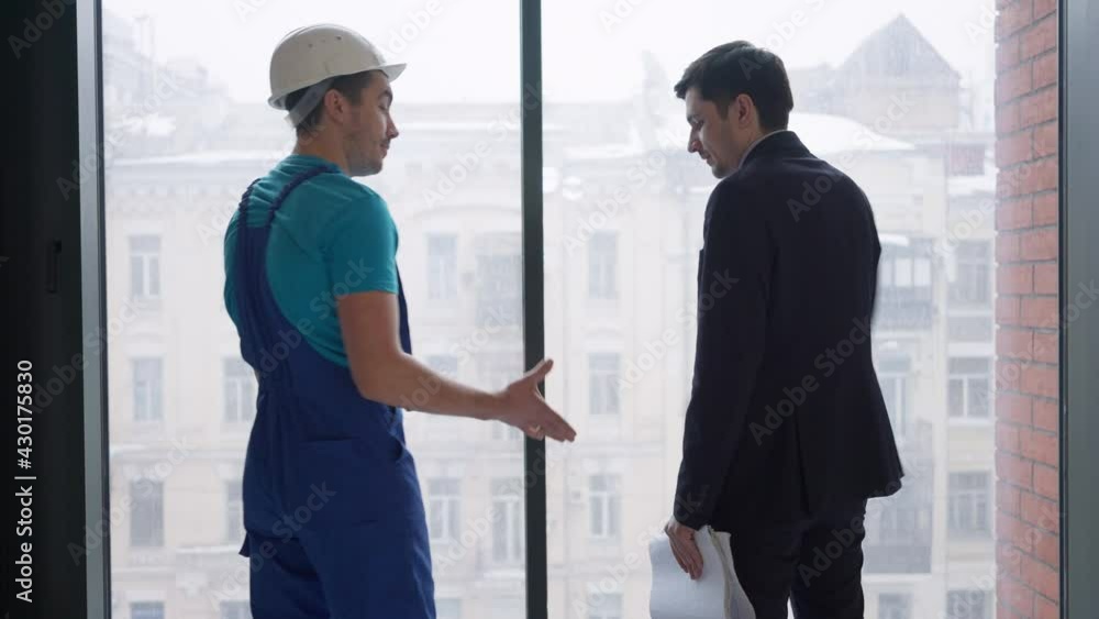 Young confident builder and buyer shaking hands standing at window indoors. Back view of professional Caucasian service man and premises owner making deal in new flat or office. Repair renovation Young confident builder and buyer shaking hands standing at window indoors. Back view of professional Caucasian service man and premises owner making deal in new flat or office. Repair renovation