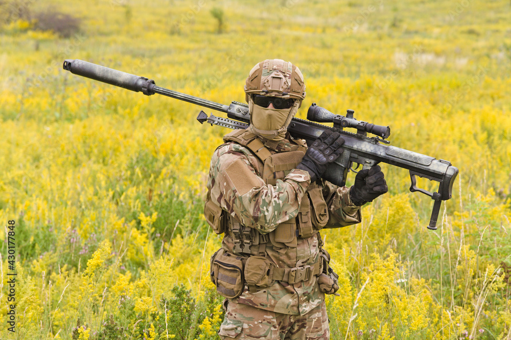 Fototapeta premium Soldier with big sniper rifle walks across blooming field