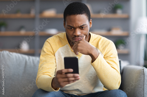 Closeup of thoughtful black guy sitting on couch, using smartphone