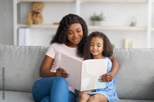 Family stay home pastimes. African American mother and her little daughter reading book together in living room