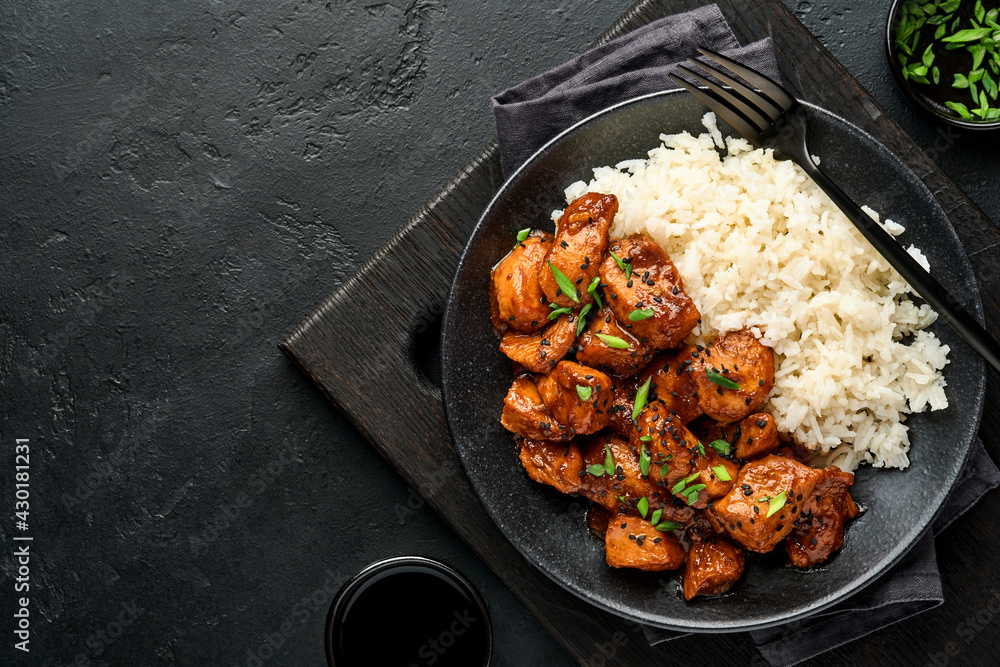 Spicy teriyaki chicken fillet pieces with rice, green onions and black sesame seeds on black plate on a dark slate, stone or concrete background. Top view with copy space.