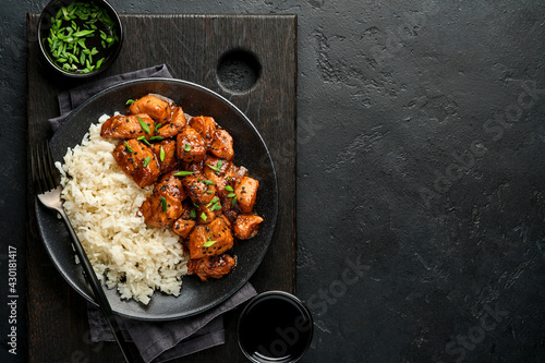 Spicy teriyaki chicken fillet pieces with rice, green onions and black sesame seeds on black plate on a dark slate, stone or concrete background. Top view with copy space.