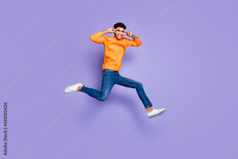 Photo of sweet cute dark skin guy dressed orange pullover jumping running v-signs cover eyes isolated purple color background