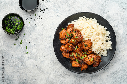 Spicy teriyaki chicken fillet pieces with rice, green onions and black sesame seeds on black plate on a grey slate, stone or concrete background. Top view with copy space.