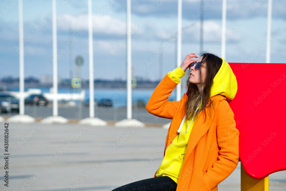 Fototapeta premium Millennial Hipster Girl in lilac sunglasses, orange coat, yellow hoodie posing at urban background. Trendy colorful casual outfit. Street Style. Fashion lookbook concept