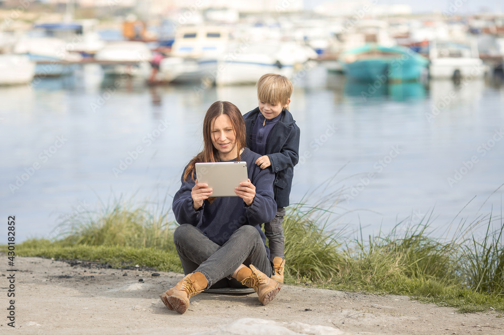 a woman with a child sitting on the beach, they communicate with someone via video communication via a tablet. Maybe they're on a trip . The concept of family, the love of the son and mother. a woman with a child sitting on the beach, they communicate with someone via video communication via a tablet. Maybe they're on a trip . The concept of family, the love of the son and mother.