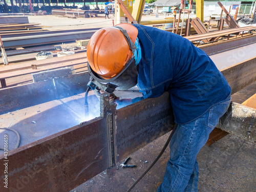 The welder is welding add joint of steel H-beam with process Flux Cored Arc Welding(FCAW) and dressed properly with personal protective equipment(PPE) for safety, at industrial factory.