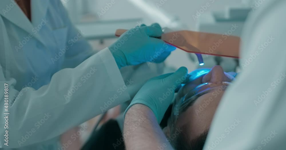 Close-up an Arabian male patient who is laying in the dental chair with a mouth opener and disposable eyeglasses is having ultraviolet teeth whitening procedure. Close-up an Arabian male patient who is laying in the dental chair with a mouth opener and disposable eyeglasses is having ultraviolet teeth whitening procedure.