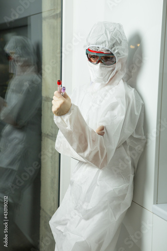 nurse wearing protection suite, gloves, breather and protective glass stands near closet with glass-tubes in her hands photo