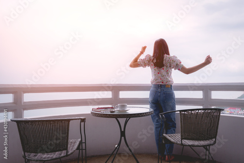Rear view of woman resting and stretching lazily after finished work beside are coffee cup, notebook, car key at hotel balcony, background is the sea. Vacation holiday relaxing concept.