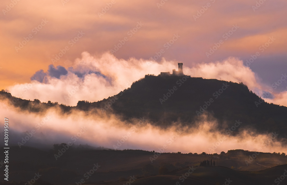 Rocca di Radicofani main tower fortification covered with morning mist. Sunrise light covering the curly clouds running over meadows and fields making light-shadows playing on Tuscany hills landscape.