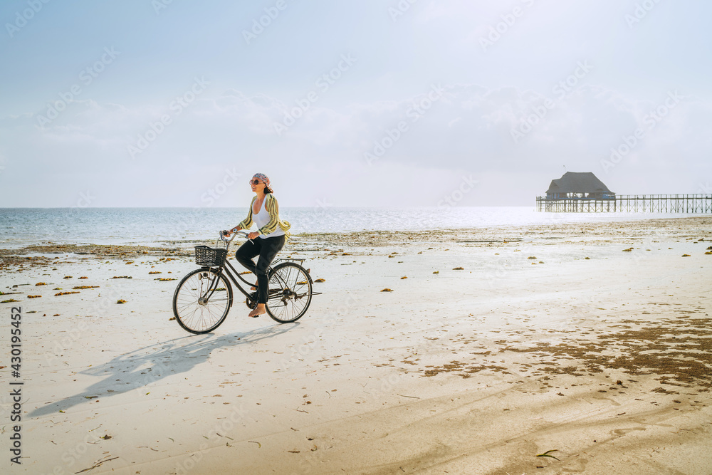 Obraz premium Young woman dressed light summer clothes riding old vintage bicycle with front basket on the lonely low tide ocean white sand coast on Kiwengwa beach on Zanzibar island, Tanzania.