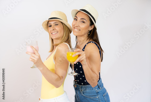 Two cheerful young women girls friends in summer clothes with straw hat, holding beverages isolated over white background.