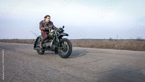Side view of a stylish cool young man leather jacket driving his vintage motorcycle on an asphalt road at late autumn cloudy evening.