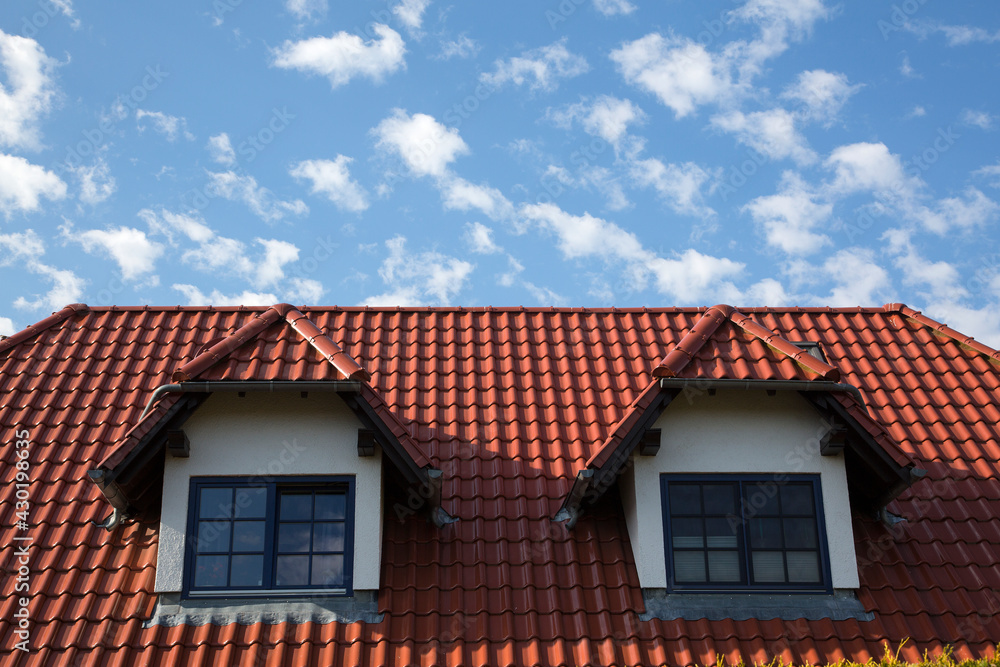 Hausdach mit zwei Dachgauben und roten Ziegeln vor einem blauen Himmel mit Wölkchen