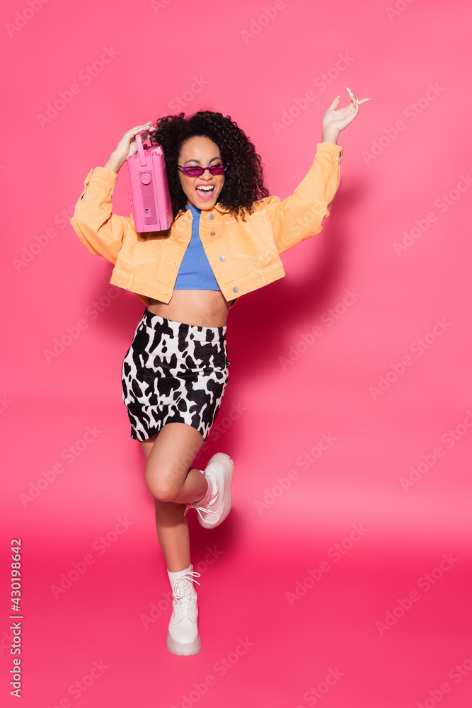 full length of happy african american woman in sunglasses holding boombox and posing on pink full length of happy african american woman in sunglasses holding boombox and posing on pink