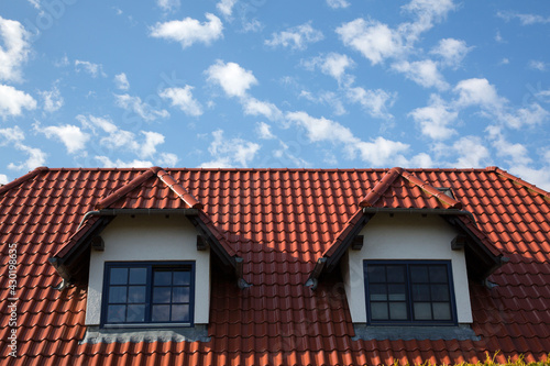 Hausdach mit zwei Dachgauben und roten Ziegeln vor einem blauen Himmel mit Wölkchen