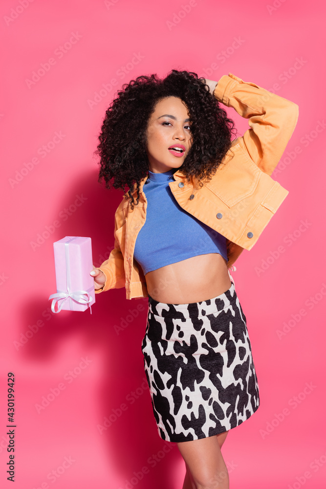 curly african american woman in crop top, skirt and jacket holding present on pink curly african american woman in crop top, skirt and jacket holding present on pink