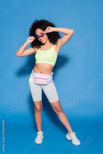 full length of happy african american woman with waist bag adjusting sunglasses on blue