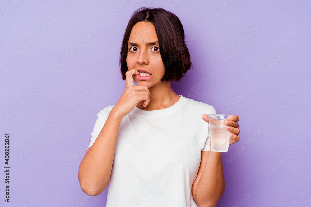 Young mixed race holding a glass of water isolated on purple background biting fingernails, nervous and very anxious. Young mixed race holding a glass of water isolated on purple background biting fingernails, nervous and very anxious.