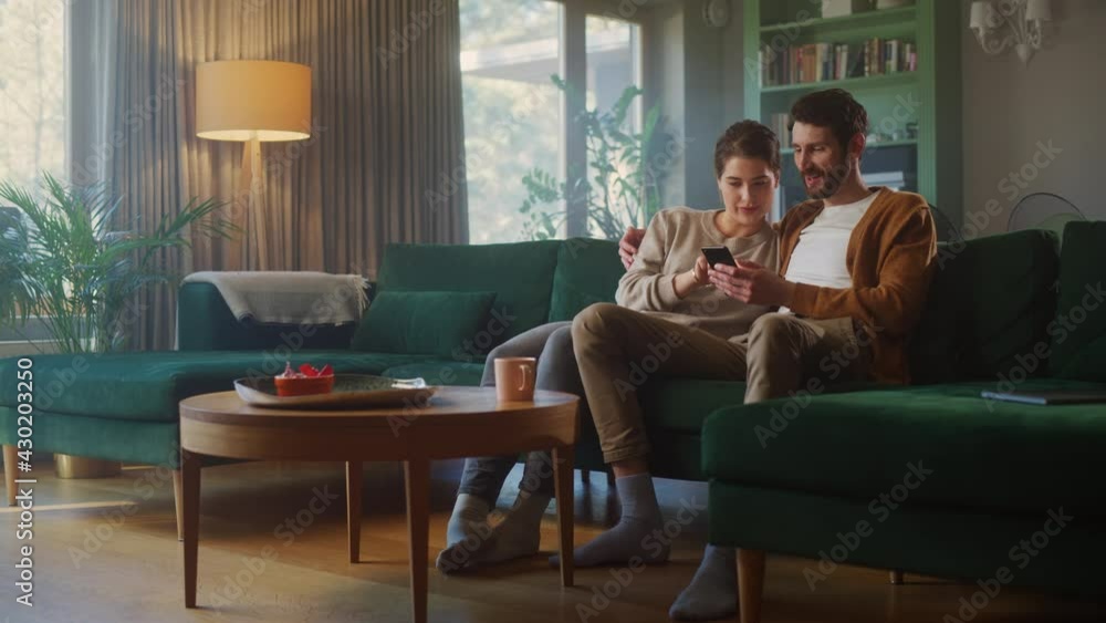 Couple Use Smartphone Device, while Sitting on a Couch in the Cozy Apartment. Boyfriend and Girlfriend Talk, do e-Shopping on Internet, Watching Funny Videos, Use Social Media, Streaming Service. Couple Use Smartphone Device, while Sitting on a Couch in the Cozy Apartment. Boyfriend and Girlfriend Talk, do e-Shopping on Internet, Watching Funny Videos, Use Social Media, Streaming Service.
