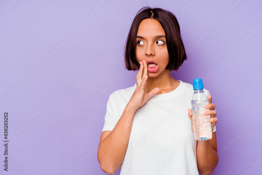 Young mixed race holding a water bottle isolated on purple background is saying a secret hot braking news and looking aside Young mixed race holding a water bottle isolated on purple background is saying a secret hot braking news and looking aside