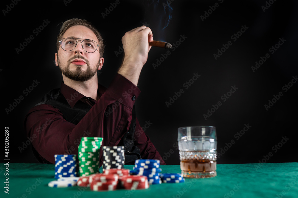 A young poker player smokes a cigar and plays poker. He has quality whiskey and a lot of chips on the table. A young poker player smokes a cigar and plays poker. He has quality whiskey and a lot of chips on the table.