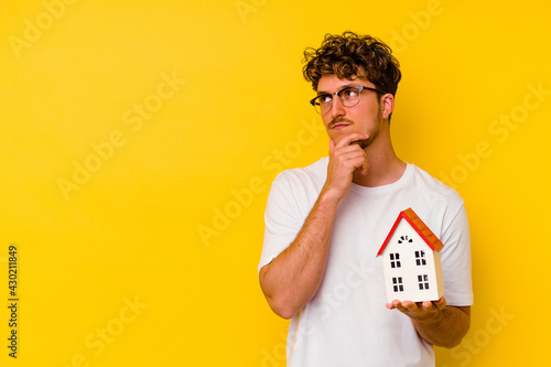 Foto Young caucasian man holding a model house isolated on yellow background looking sideways with doubtful and skeptical expression