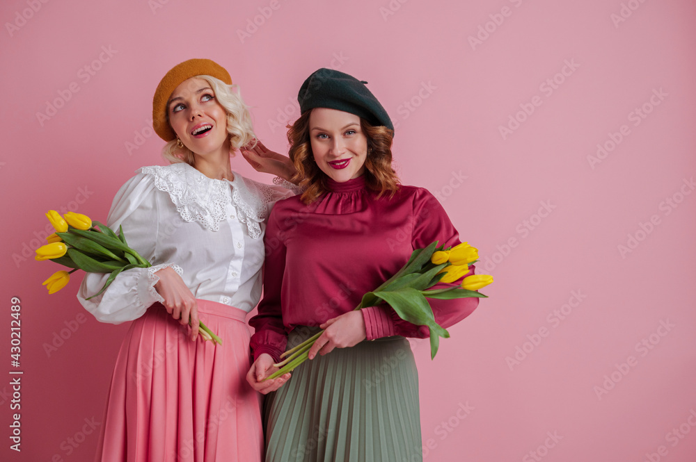 Two happy smiling dreamful woman, friends, wearing colorful shirts, berets, skirts, holding tulips, posing on pink background. Spring fashion, Women's Day celebration, advertising concept

