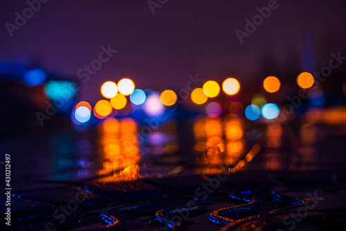 Rainy night in the city. Parking mall. Reflections of shop windows in a puddle on asphalt. Colorful colors. Close up view of a hatch at the level of the pavement.