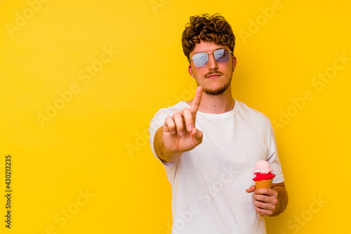 Young caucasian man eating an ice cream isolated on yellow background showing number one with finger. © Asier