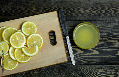 slices of fresh juicy bright yellow lemons laid out on a light wooden cutting board and a glass plate with lemon juice and a knife on a dark wooden table, taken close-up