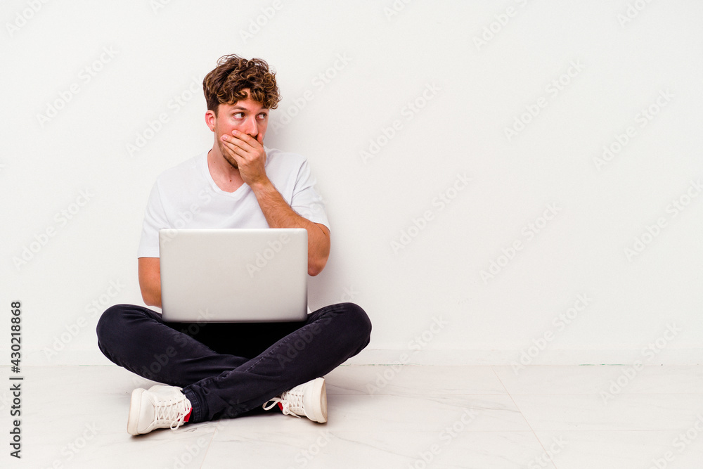 Young caucasian man sitting on the floor holding on laptop isolated on white background thoughtful looking to a copy space covering mouth with hand. Young caucasian man sitting on the floor holding on laptop isolated on white background thoughtful looking to a copy space covering mouth with hand.