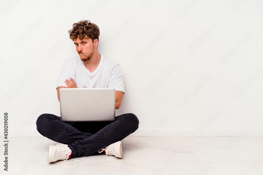 Young caucasian man sitting on the floor holding on laptop isolated on white background frowning face in displeasure, keeps arms folded. Young caucasian man sitting on the floor holding on laptop isolated on white background frowning face in displeasure, keeps arms folded.