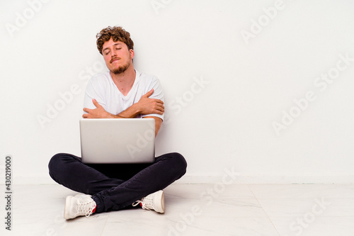 Young caucasian man sitting on the floor holding on laptop isolated on white background hugs, smiling carefree and happy.
