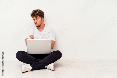 Young caucasian man sitting on the floor holding on laptop isolated on white background frowning face in displeasure, keeps arms folded.