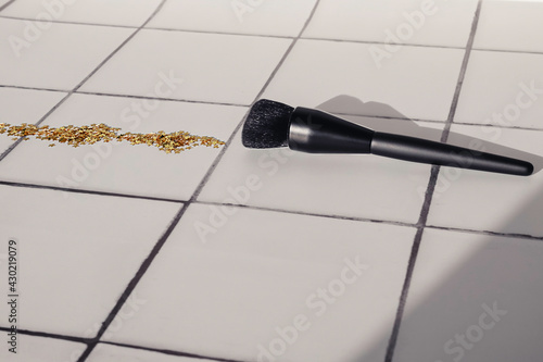 A black makeup brush for powder or foundation and golden glitter sequins on white tile floor or countertop background.