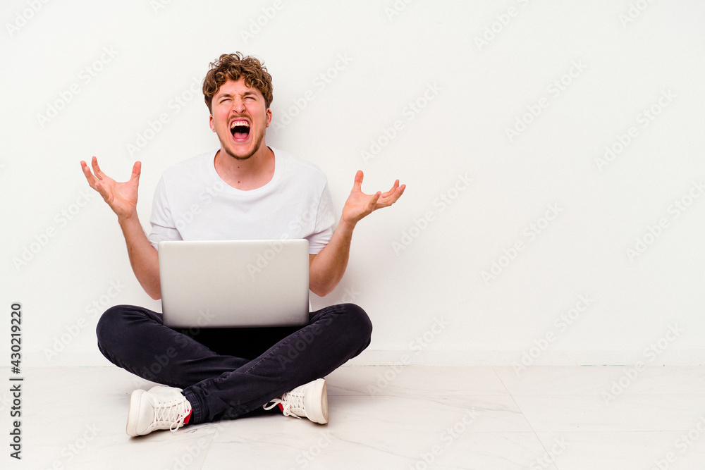Young caucasian man sitting on the floor holding on laptop isolated on white background screaming with rage. Young caucasian man sitting on the floor holding on laptop isolated on white background screaming with rage.