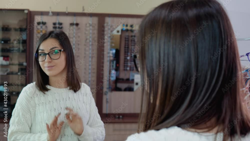 Young brunette with blue eyes in a cozy white sweater trying on glasses in front of a mirror in an optics store Young brunette with blue eyes in a cozy white sweater trying on glasses in front of a mirror in an optics store