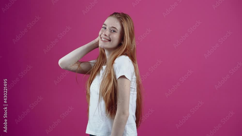The young redhead, charismatic looks dreamily up with a wide smile, playfully puts her hand on the ends of her hair. Freckled teenage girl in a white T-shirt, isolated on a pink background. The The young redhead, charismatic looks dreamily up with a wide smile, playfully puts her hand on the ends of her hair. Freckled teenage girl in a white T-shirt, isolated on a pink background. The