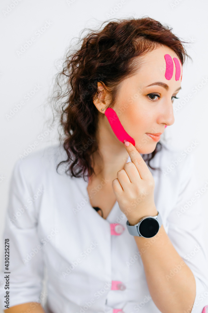 a dark-haired Caucasian woman in a white medical gown puts pink tapes on her face. the concept of cosmetology and health. multi-colored patch for muscles a dark-haired Caucasian woman in a white medical gown puts pink tapes on her face. the concept of cosmetology and health. multi-colored patch for muscles