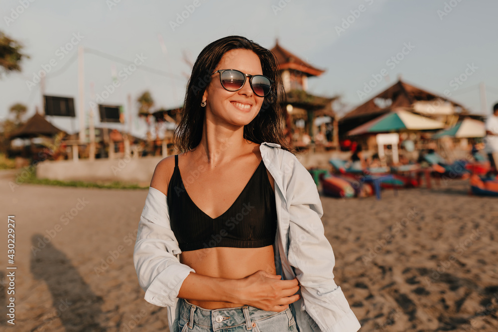 Fototapeta premium Outdoor portrait of attractive charming girl with dark hair wearing sunglasses and shirt walking down the beach in sunlight near the ocean