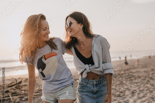 Outdoor portrait of two young pretty women having fun on sunset near the ocean on sandy beach. Two friends walking down the beach in warm day