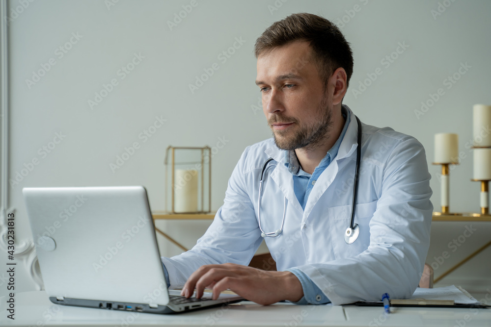 Concentrated doctor working with laptop at desk in office. Male doctor using laptop at desk in clinic Concentrated doctor working with laptop at desk in office. Male doctor using laptop at desk in clinic