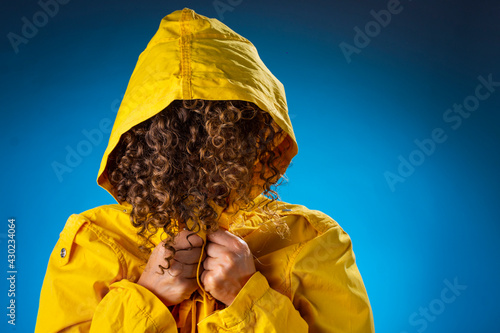 Mujer joven oculta su rostro con cabello enrulado con impermeable amarillo sobre fondo azul