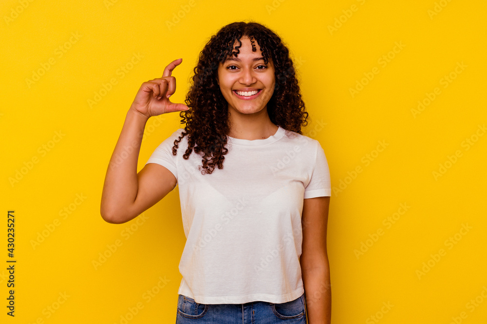 Young mixed race woman isolated on yellow background holding something little with forefingers, smiling and confident. Young mixed race woman isolated on yellow background holding something little with forefingers, smiling and confident.