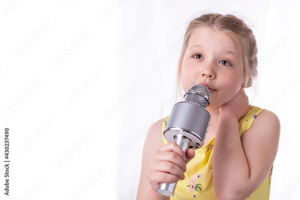 Fototapeta premium A blonde little girl sings a song into a microphone in her hands. A beautiful child shows his sincere emotions of happiness and positive. Braided hair in pigtails and a yellow dress