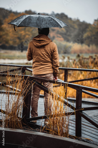 Rear view of a young man in an orange jacket, he stands and holds his black umbrella in an autumn park